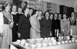 En grupp kvinnor under en v�vkurs med v�var i bakgrunden och ett dukat kaffebord framf�r sig - klicka f�r att f�rstora