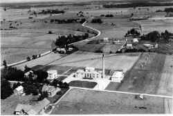 Flygfoto �ver Fjugesta by fr�n v�ster. Mitt ibild syns nya mejeriet fr�n 1938. Tomten till h�ger ovanf�r mejeriet �r den blivande H�gans hembygdsg�rd d�r loftboden och stallbyggnaden �r p� plats. Eventuellt f�rbereder man bygget av granhammarstugan. - klicka f�r att f�rstora