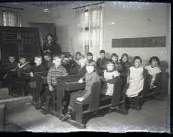 En grupp skolbarn med en lrarinna vid en svart tavla i bakgrunden. Bilden kan vara tagen i smskolan i Hackvads Via - klicka fr att frstora