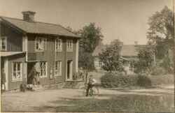 Husen på bilden ägdes av förre handlaren Axel Englund. Den närmasta byggnaden uppges ha flyttats hit från Klockaregården på 1870-talet av en handlare vid namn Nordqvist. När bilden togs hyrde handlare August Hagberg handelsboden medan veterinär Joel Ekelund bodde på övervåningen. I dörren står handlanden August Hagberg, på trappan sitter veterinär Joel Ekelund och vid cykel står snickaren och telefonföreståndaren Aron Klaesson - klicka f�r att f�rstora